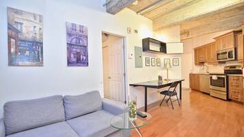 A modern apartment with a grey sofa, a glass coffee table, a dining area with a chair, and a kitchen with wooden cabinets and appliances.