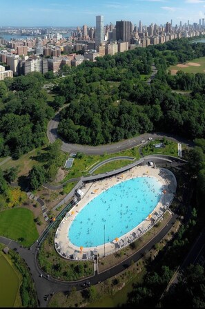 Una piscina grande y ovalada rodeada por un parque con senderos para caminar y un horizonte urbano al fondo.
