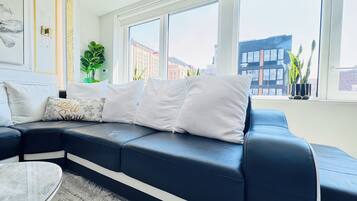 A modern living room with a black leather sofa, white cushions, a marble coffee table, and large windows offering city views.