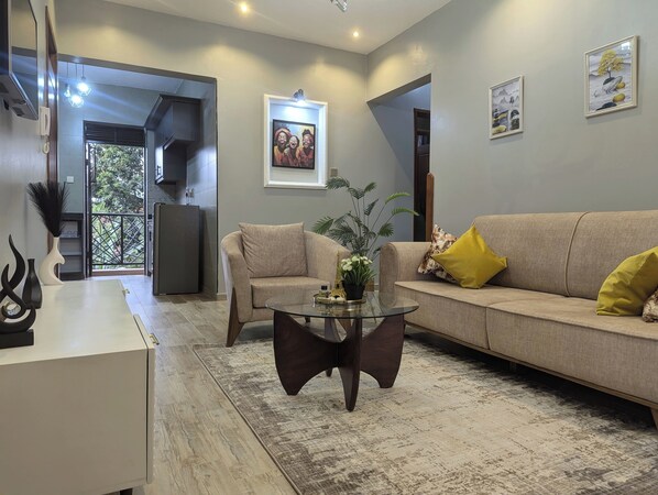 Un salon avec un canapé beige, une table basse en verre et une petite table d’appoint. On aperçoit une plante sur la table basse et un cadre photo accroché au mur.