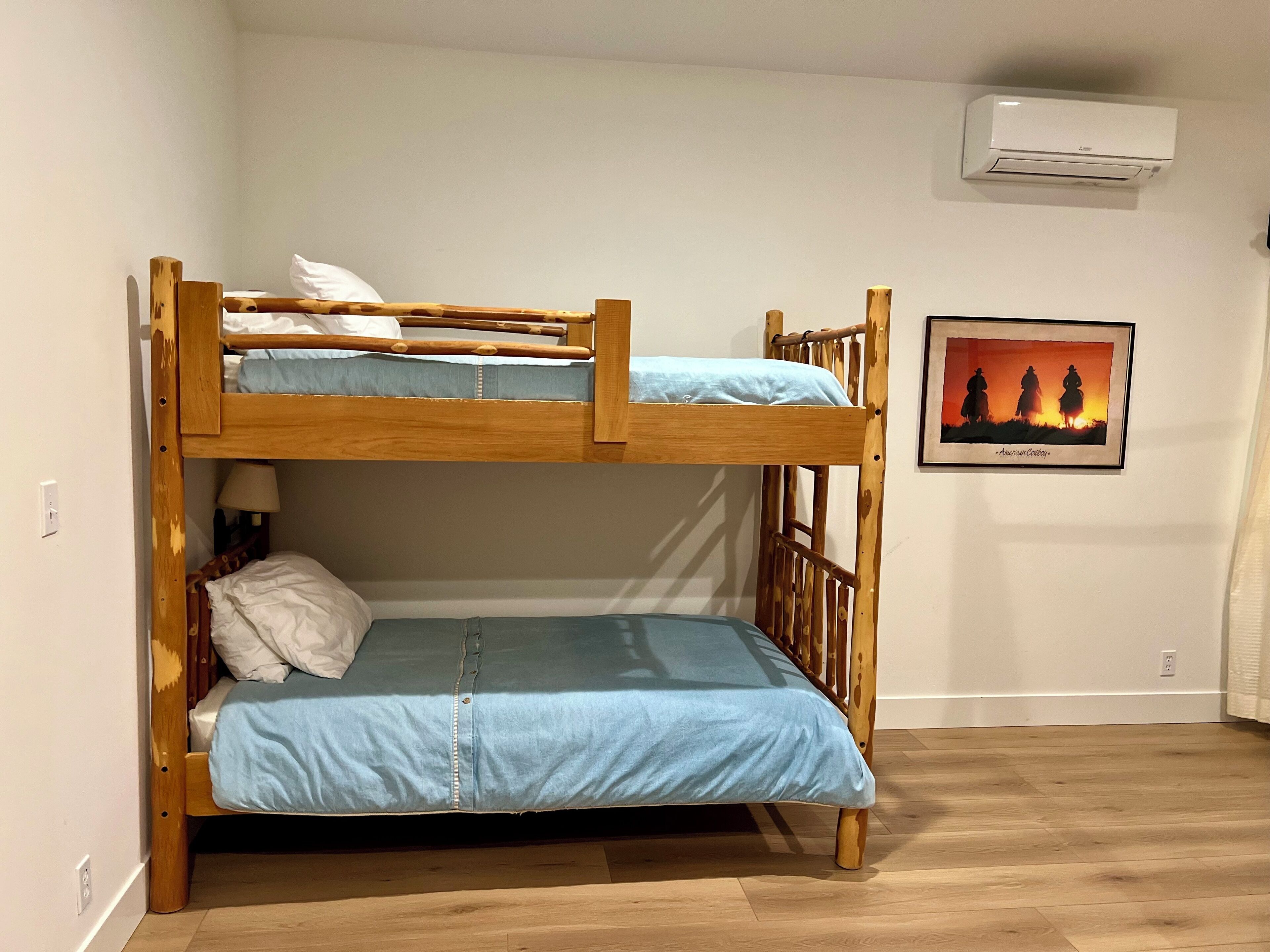 A bunk bed with a blue mattress and a wooden frame. There is a wall-mounted air conditioner above the bed and a framed picture of cowboys on horseback on the wall.