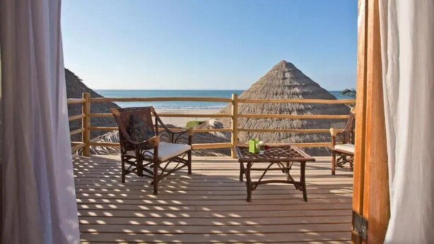 Una terraza de madera con mobiliario de mimbre y una estructura de techo de paja, con vistas a una playa y al océano.