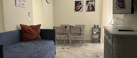 A living room with a blue sofa, a dining area with chairs, a small table with cups, and a kitchen area with a cabinet and a microwave.