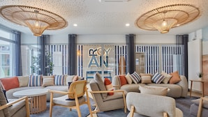 Una sala de estar moderna con un sofá seccional, sillones y una mesa de centro. Hay un ventanal con cortinas, una lámpara de techo decorativa y un letrero con el texto "ROYAL".