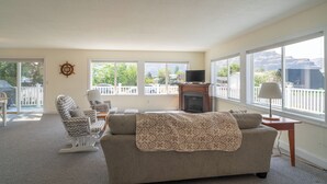 Un salon spacieux comprenant un canapé, des fauteuils, une cheminée et de grandes fenêtres donnant sur l’extérieur.