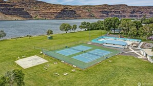Un court de tennis avec une piscine, entouré d’une pelouse et d’arbres, avec un lac et des collines en arrière-plan.