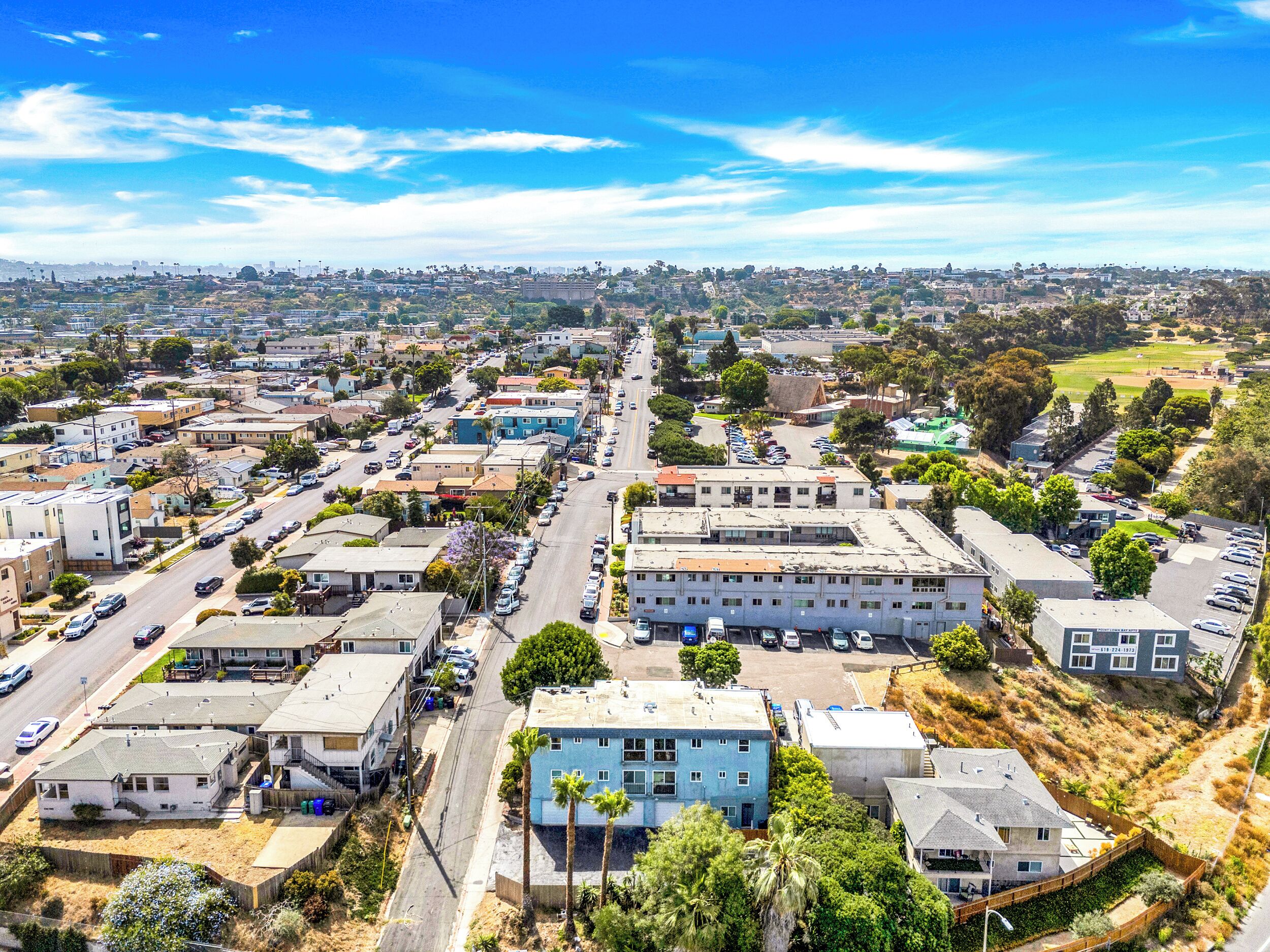 A residential area with a mix of single-family homes and small apartment buildings, featuring a central street with parked cars and a clear sky.