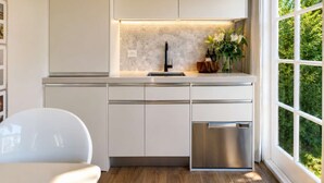 Une cuisine moderne avec des meubles blancs, un lave-vaisselle en acier inoxydable et une fenêtre donnant sur un paysage verdoyant.