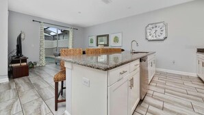 Una cocina moderna con armarios blancos, encimera de granito y un lavamanos integrado. Hay un reloj en la pared y cuadros colgados sobre la encimera.