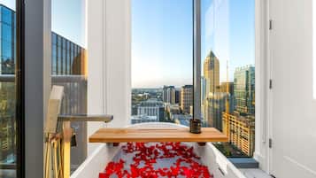 A bathtub with red rose petals, a wooden shelf, and a city view through large windows.