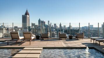 A rooftop pool area with lounge chairs and a fountain, overlooking a city skyline.