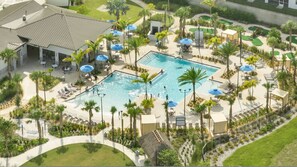 An aerial view of a resort with a central swimming pool surrounded by lounge areas, palm trees, and a golf course. - Go 31- Champions Gate (Kissimmee)