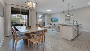 A modern kitchen with a central island, pendant lighting, and a dining area with a large table and chairs. - Go 31- Champions Gate (Kissimmee)