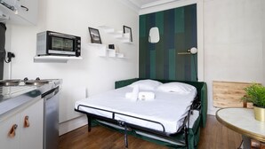 Una habitación de hotel compacta con cama, kitchenette pequeño, microondas y mesita de noche con planta.