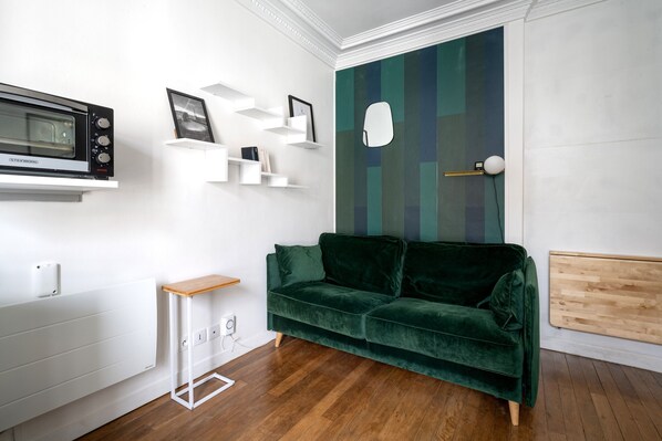 Un salon moderne avec un canapé en velours vert, des étagères blanches, un four à micro-ondes encastré et un plancher en bois.