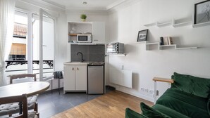 Una cocina compacta con armarios blancos, un microondas y un refrigerador pequeño. Hay una mesa de comedor con sillas y un sofá verde en la sala de estar.