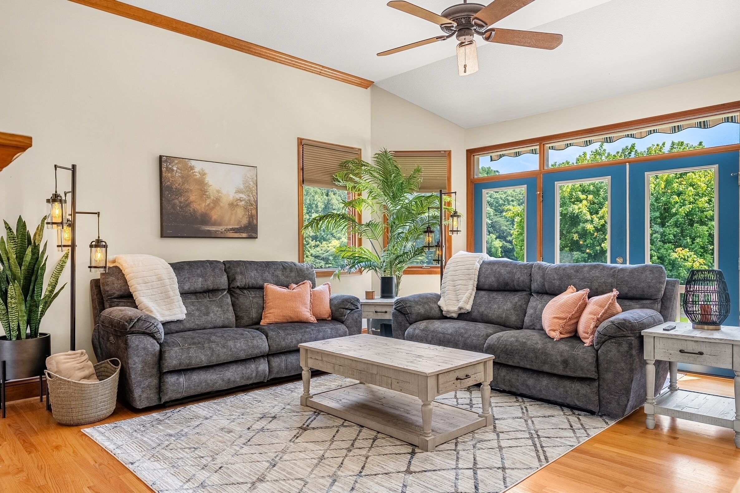 A living room with two grey sofas, a wooden coffee table, a ceiling fan, and large windows with a view of greenery.