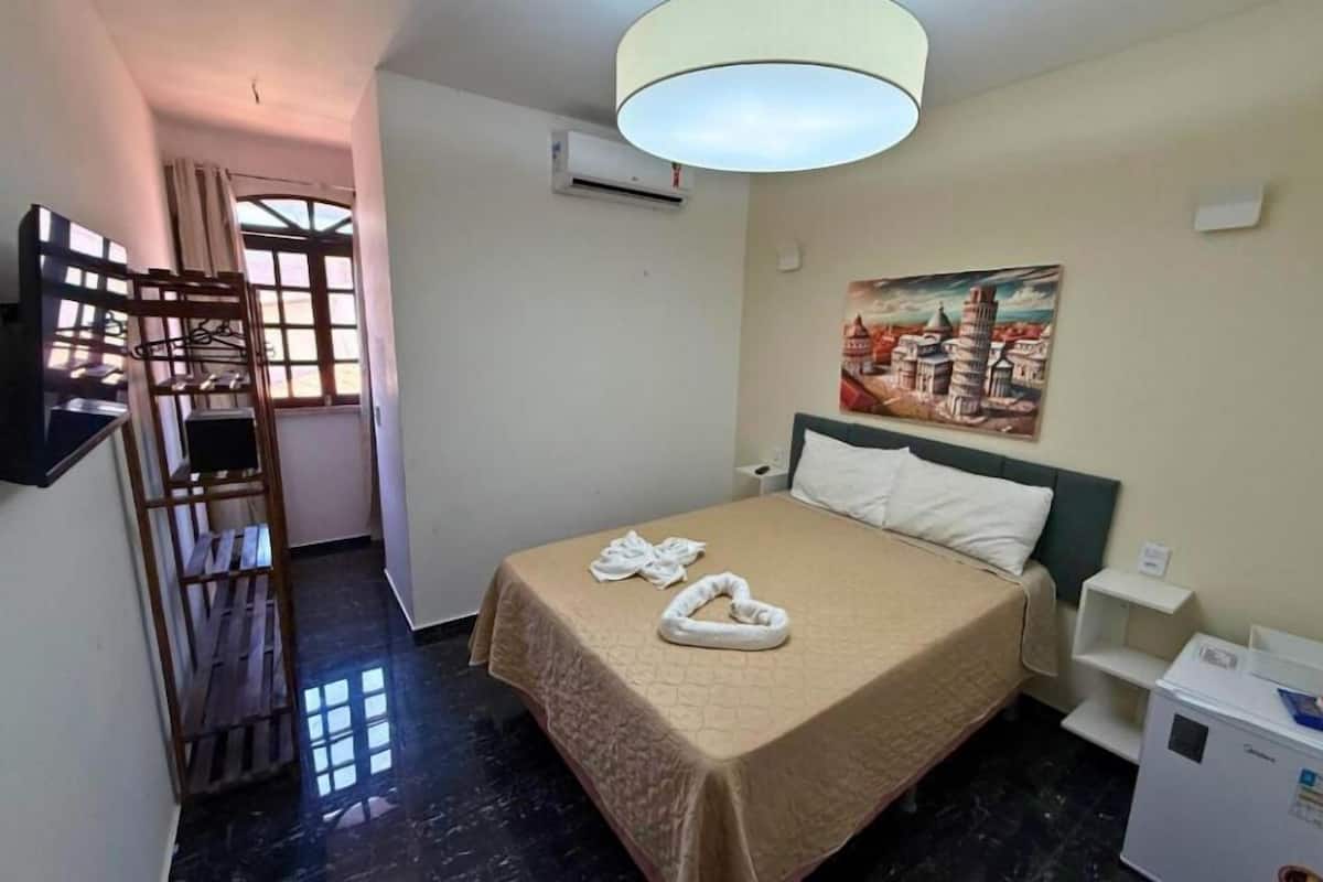 A hotel room with a bed, a painting of a cityscape, a wardrobe, and a desk with a lamp.