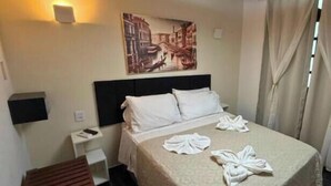A hotel room with a bed, a headboard, a wall-mounted light, a painting, a bedside table, a bench, and a curtain.