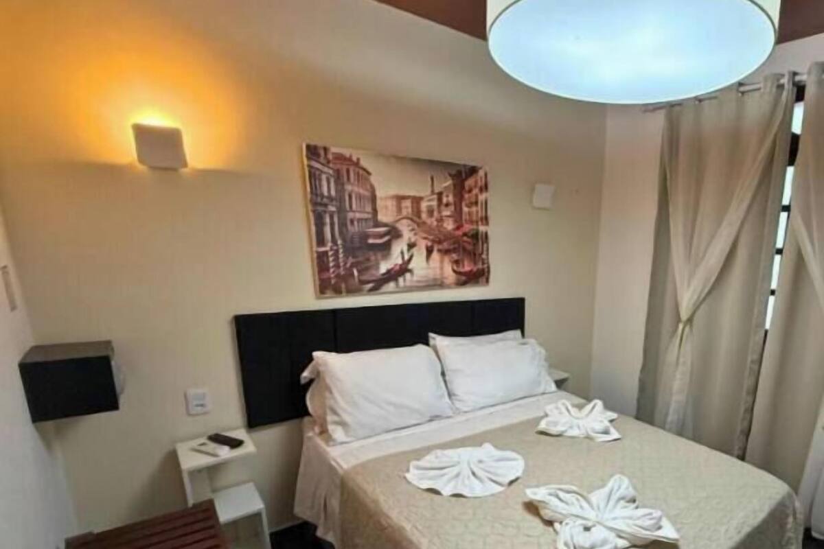 A hotel room with a bed, a headboard, a wall-mounted light, a painting, a bedside table, a bench, and a curtain.