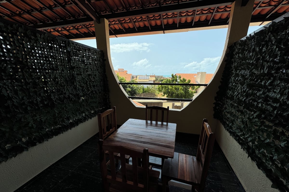 A covered patio with a table and chairs, surrounded by walls covered in foliage, and a view of the cityscape through a large window.