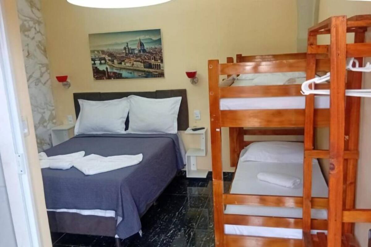 A hotel room with a bed, a wooden bunk bed, a nightstand, a wall-mounted lamp, and a painting.