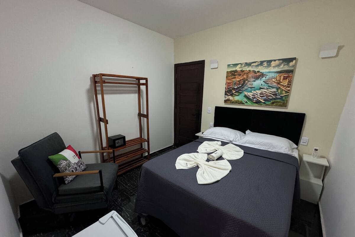 A hotel room with a bed, a chair, a wooden rack, a painting, and a small table with a phone.