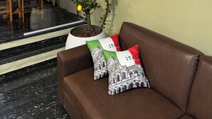A living room with a brown leather sofa, two pillows with Italian designs, a potted lemon tree, and a glass partition with a wooden chair and table in the background.