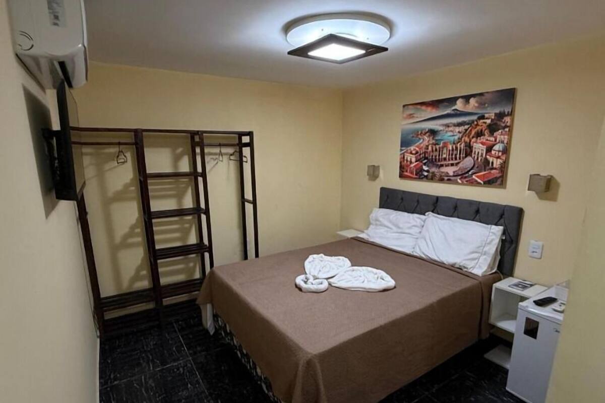 A hotel room with a bed, a nightstand, a small table, a wall-mounted air conditioner, a painting, and a wardrobe with a mirror.