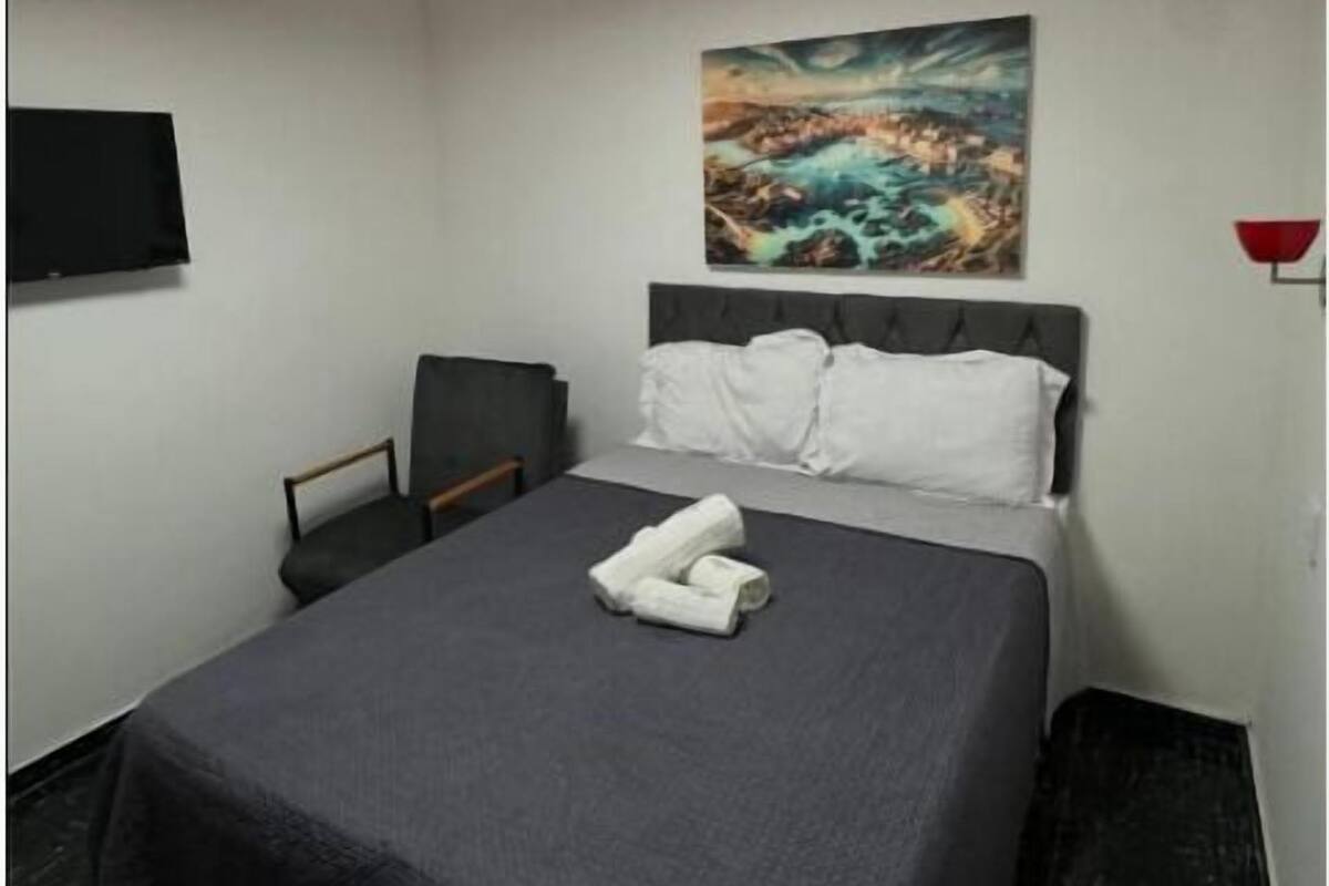 A hotel room with a bed, a painting on the wall, a flat-screen TV, and a red ceiling light.