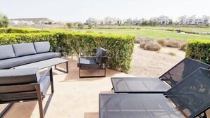 Ein Sitzbereich im Freien mit einer Couch und Sesseln, einem Glastisch und Blick auf einen angelegten Garten sowie Gebäude im Hintergrund.