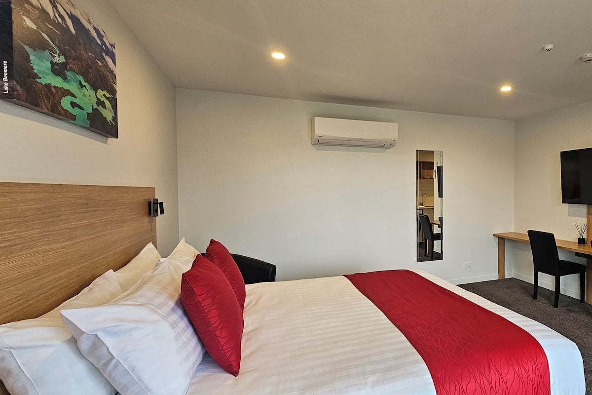 A hotel room with a bed, a desk, a chair, a TV, and an air conditioning unit.