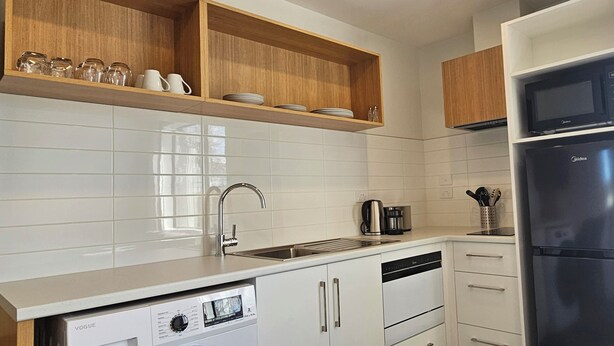 A modern kitchen with white cabinetry, a built-in dishwasher, a stainless steel refrigerator, and wooden shelves above the sink. - Alps Motel Apartment Twizel (Twizel)