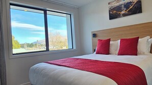 A hotel room with a large bed, red and white bedding, a wooden headboard, a window with a view of trees and mountains, and a painting on the wall. - Alps Motel Apartment Twizel (Twizel)
