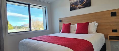 Una habitación de hotel con una cama grande, ropa de cama roja y blanca, un cabecero de madera, una ventana con vistas a árboles y montañas, y un cuadro en la pared.