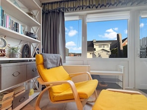 A room with a yellow armchair, a bookshelf with books, a clock, and a view of buildings through large windows.