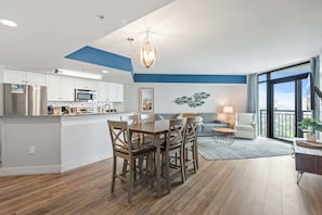A modern kitchen with stainless steel appliances, a dining area with wooden chairs, and a living room with a sofa and a large window.