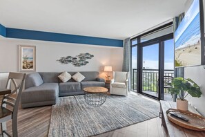 Ein modernes Wohnzimmer mit einem grauen Sofa, einem runden Couchtisch und einem großen Fenster mit Blick auf den Strand.