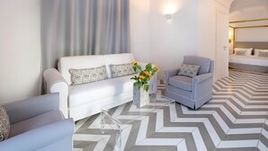 A modern living room with a white sofa, a grey armchair, and a coffee table with a vase of flowers. The room has a geometric patterned floor and a bed in the background.