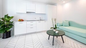A modern kitchen with white cabinets, a sink, and a countertop with fruit. There's a green plant and a round table with a vase of flowers. A light blue sofa is against the wall.