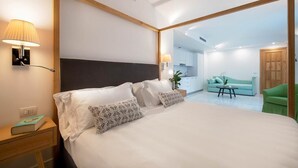 A hotel room with a large bed, bedside lamps, a small table, a green armchair, and a view into the kitchen area.