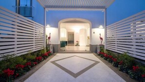 A covered entrance with a geometric tile floor, flanked by white slatted structures and red flowers.