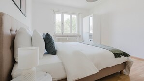 Ein Schlafzimmer mit einem großen Bett, einem Fenster, einem Heizkörper und einer Tür zu einem anderen Raum.