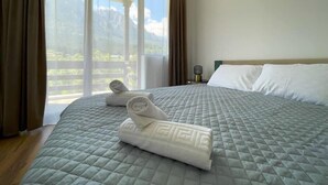 A hotel room with a large bed, a nightstand, and a view of mountains through the window. - Alpine River Home: Peace and Mountain View (Bușteni)