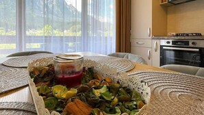 A dining table with a centerpiece of green leaves and a jar of red liquid, a kitchen with a stove and oven, and a view of mountains through large windows. - Alpine River Home: Peace and Mountain View (Bușteni)