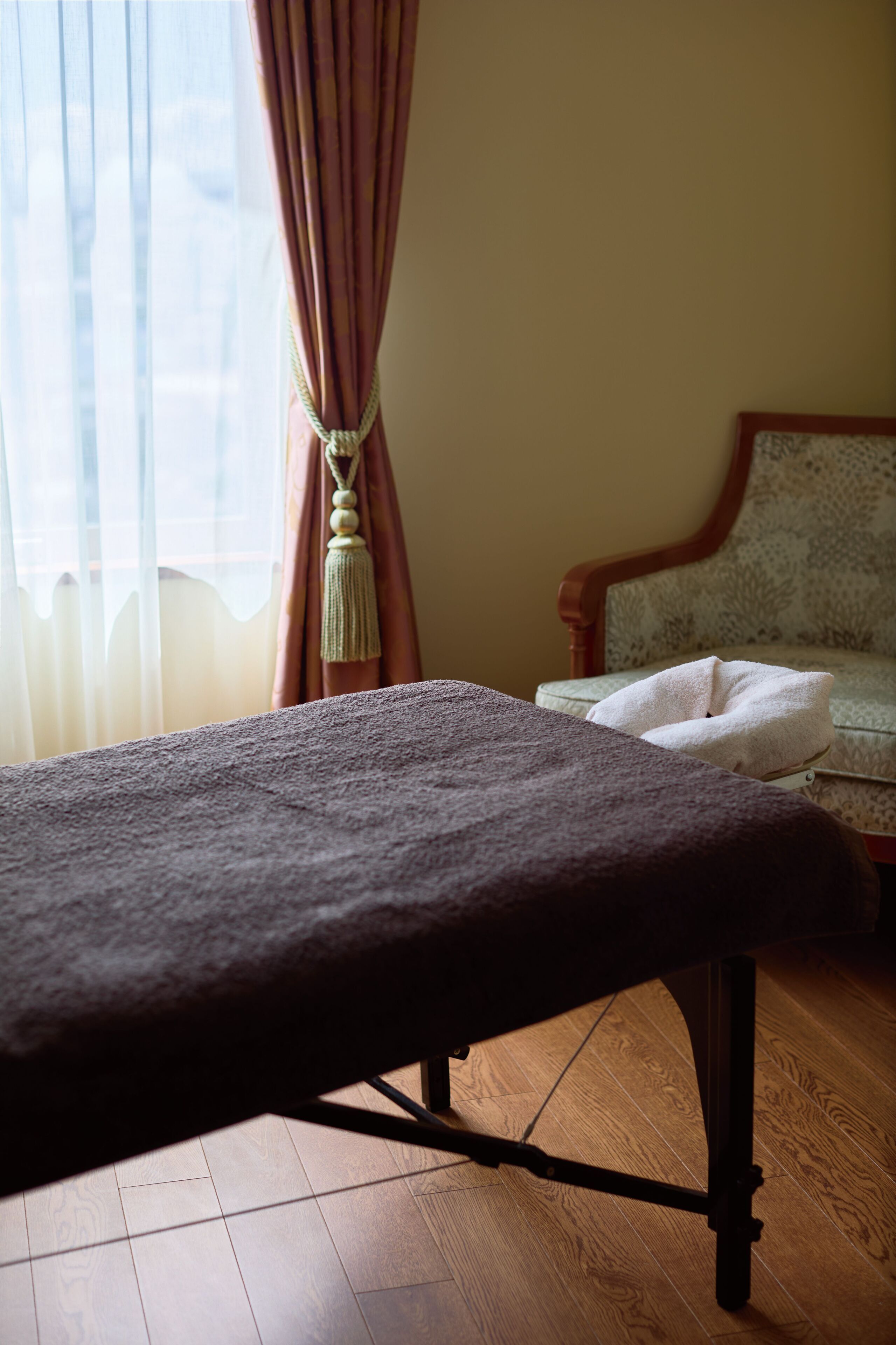 A room with a wooden floor, a bed with a dark blanket, a chair with a floral pattern, and a window with curtains.