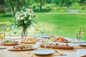 A table set for a meal with various dishes and glasses of wine, surrounded by a lush green garden.