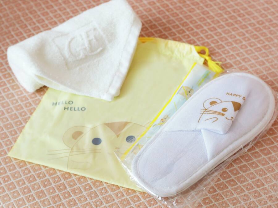 Una toalla blanca doblada con cuidado, un paño amarillo con un diseño de gato, y un par de pantuflas blancas con estampado de gato.