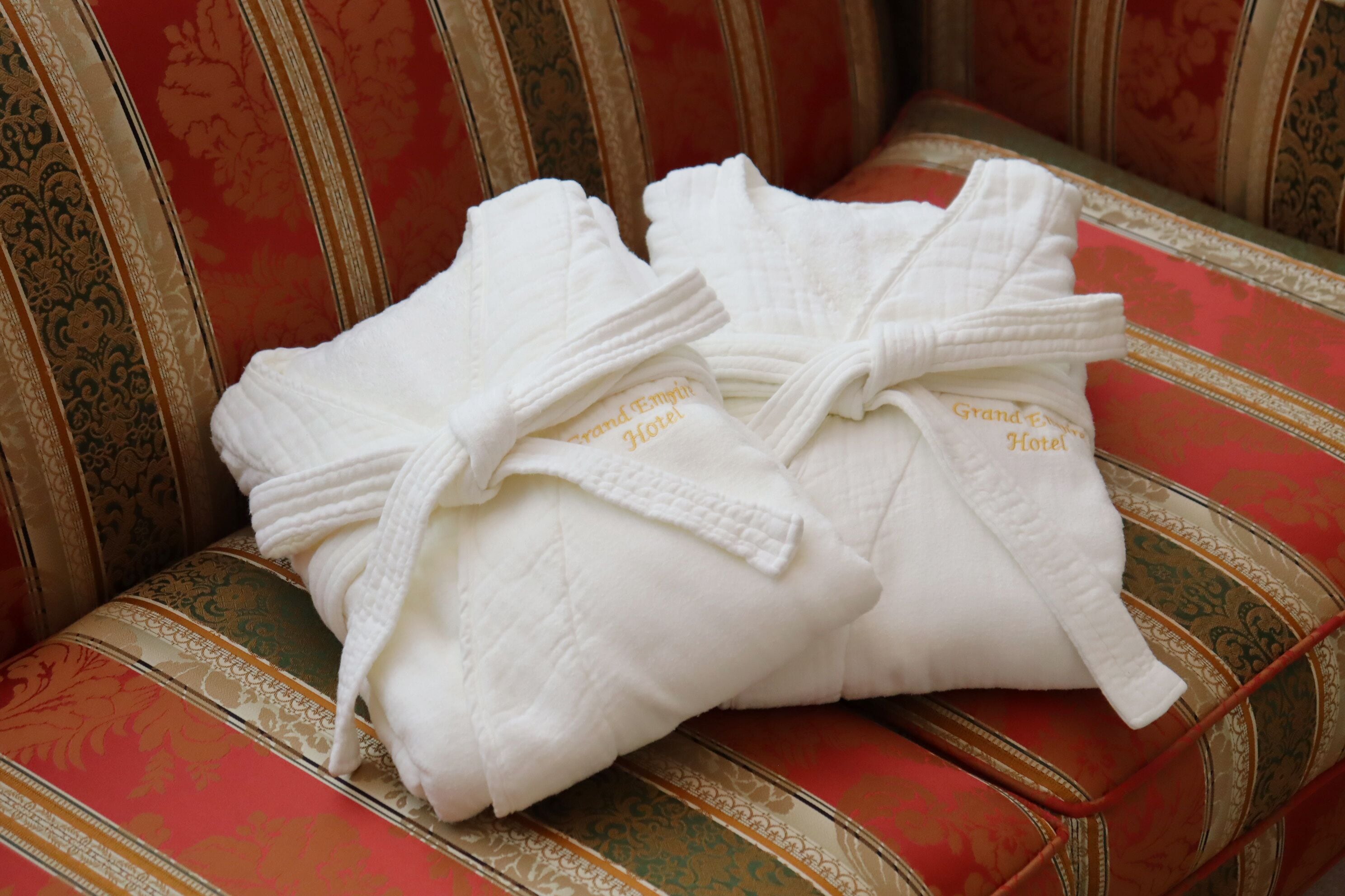 Duas camisas brancas com a inscrição 'Grand Emporio Hotel' bordada, colocadas em um sofá com padrão em vermelho e dourado.