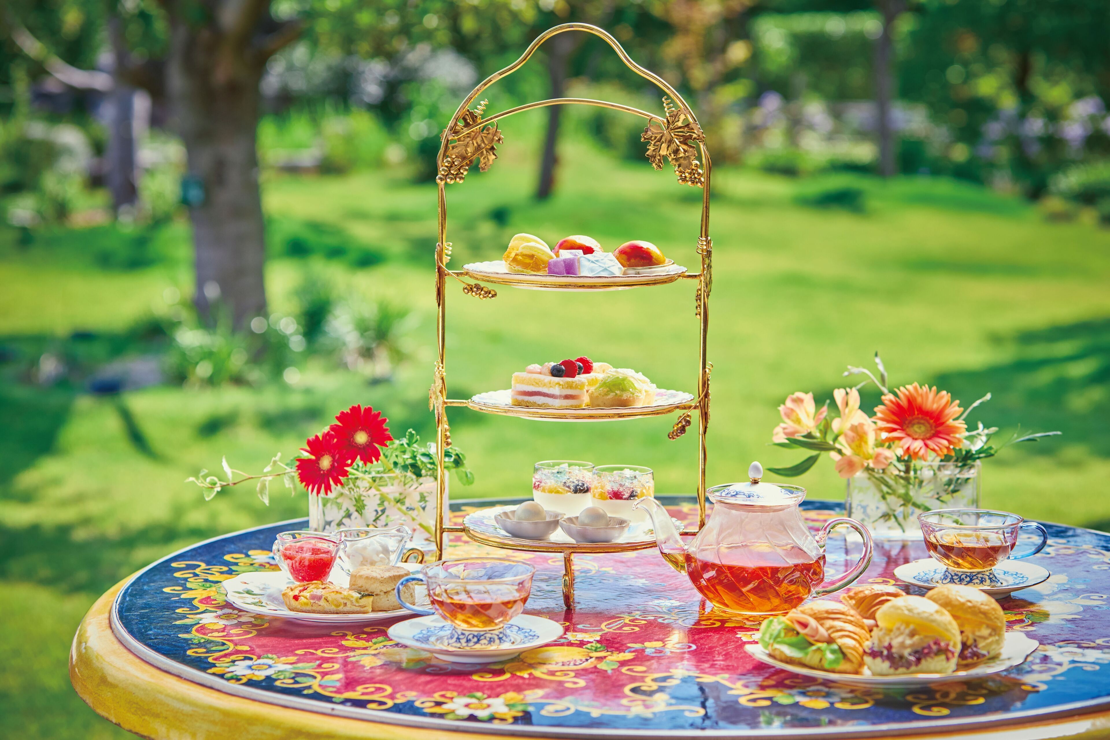 A three-tiered tea service stand with various pastries and a floral arrangement on a round table outdoors.